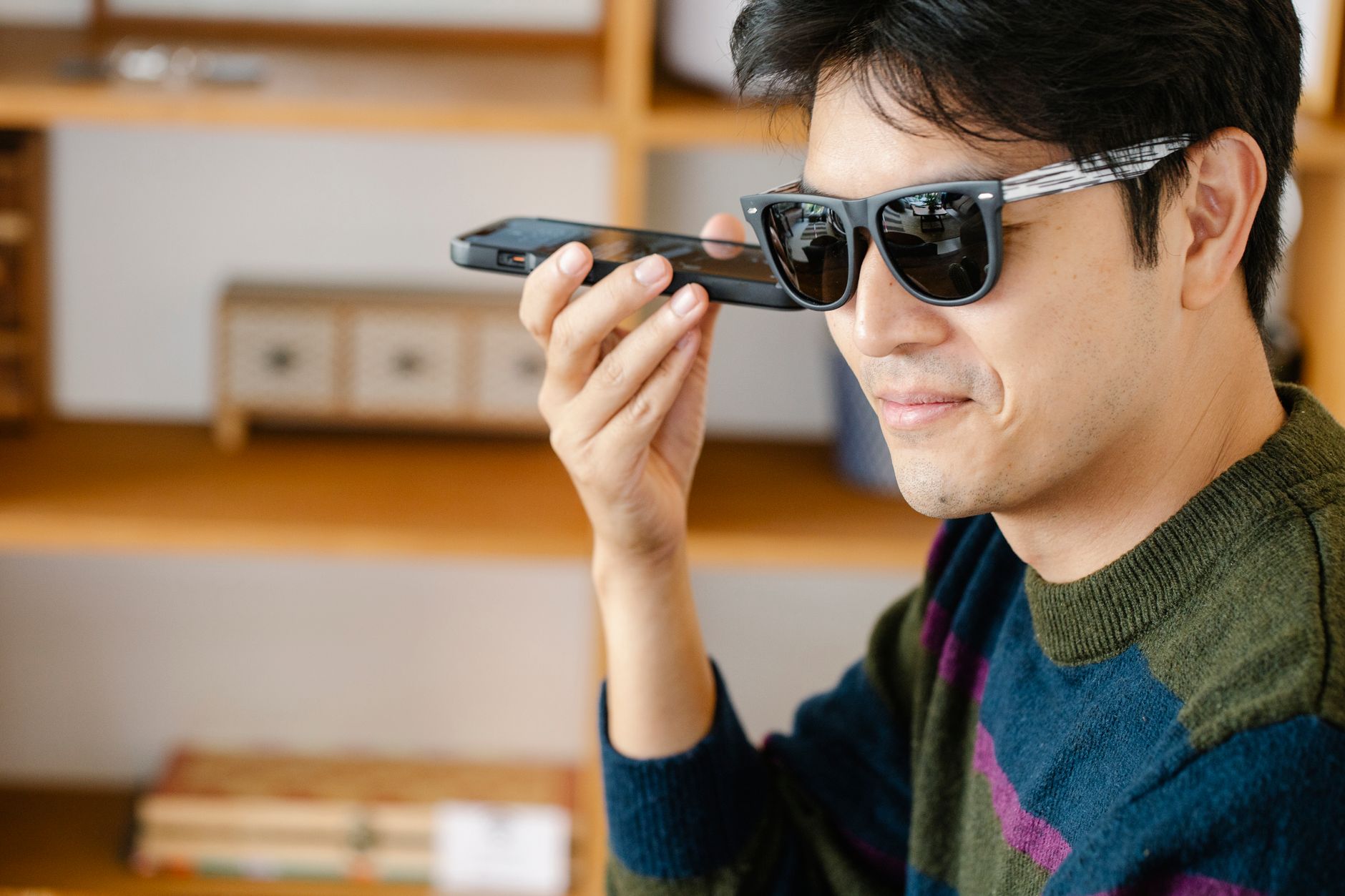 A cheerful man wearing sunglasses indoors, using voice commands on his smartphone.