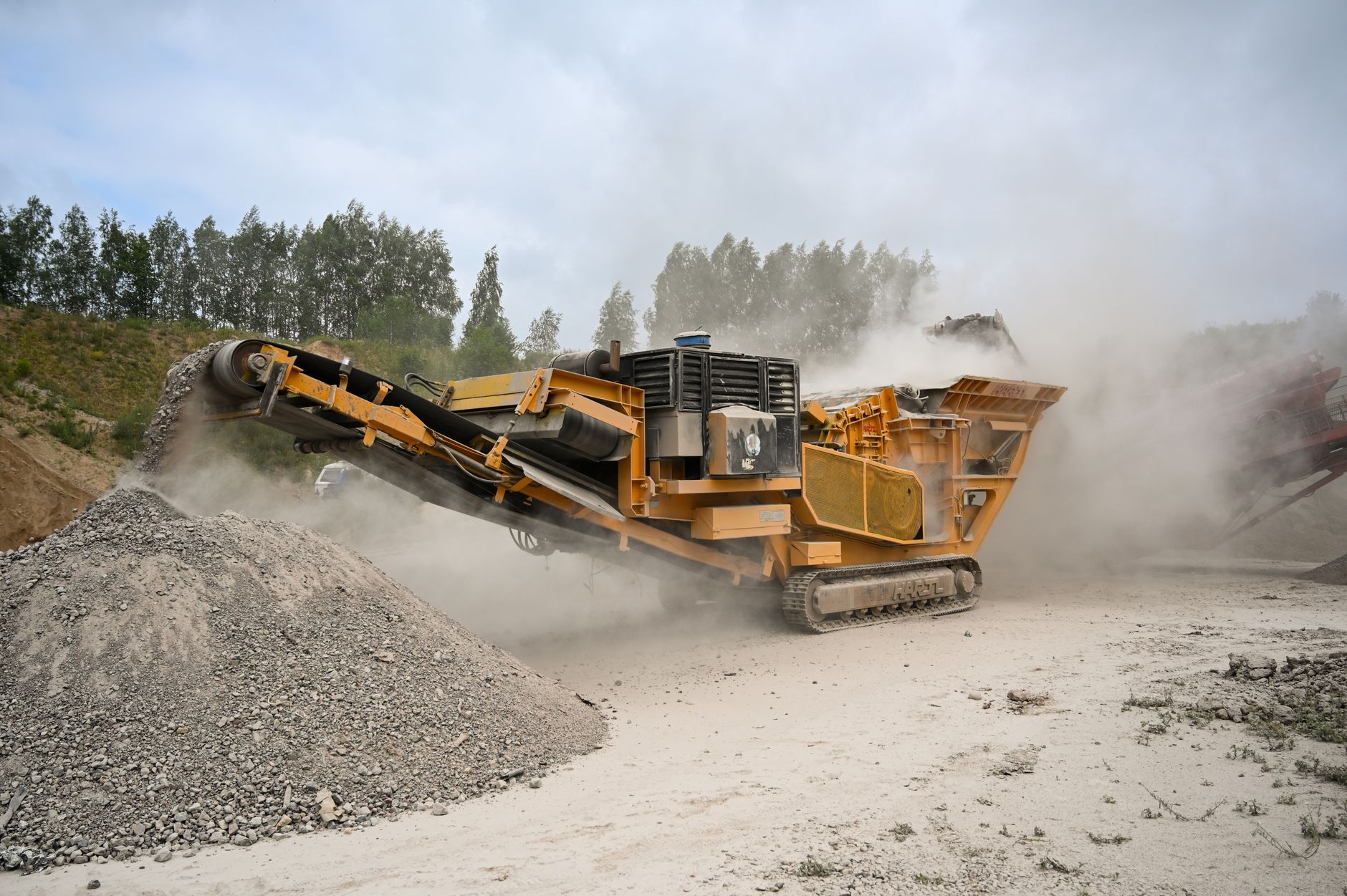 Heavy-duty crusher machine processes materials in dusty quarry amidst greenery.